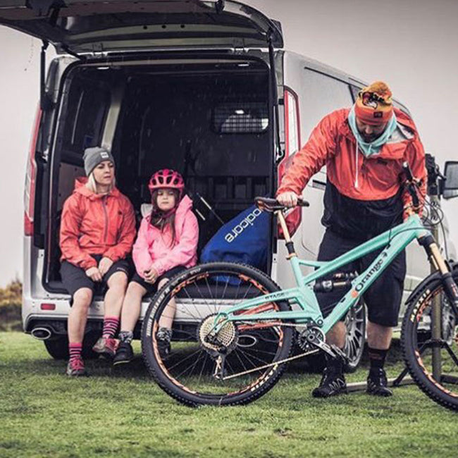 lady and child sat in boot of van with booicore dirtbag royal blue next to them, man in front holding mountain bike