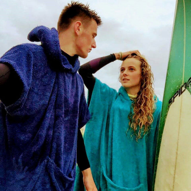 man wearing booicore changing robe navy blue next to lady wearing booicore changing robe turquoise, holding a surfboard